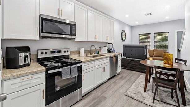 A modern kitchen with white cabinets, a microwave, stove, and dishwasher. There's a dining area with a table and chairs, and a living room with a TV and sofa. - Cozy 2bdr/1br Home near downtown (Sacramento)