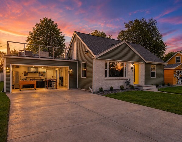 A two-story house with a garage, a patio, and a well-lit interior. - Lambeau View 🏈 | Pool · Spa 💦 | Rooftop · Fire Table 🔥 | Entertainment Room 🎉 (Green Bay)