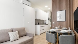 Uma sala de estar moderna com um sofá, uma mesa de jantar para dois e uma cozinha ao fundo.