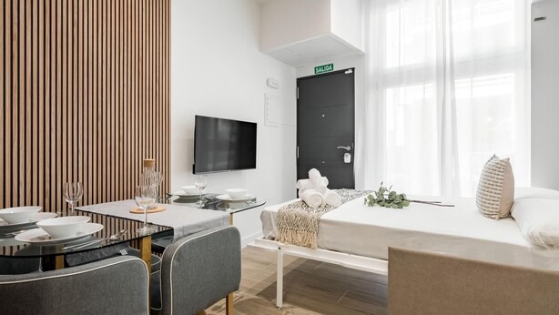 Quarto de hotel moderno com área de refeições, televisão de tela plana, cama com edredom branco e uma grande janela com cortinas leves.
