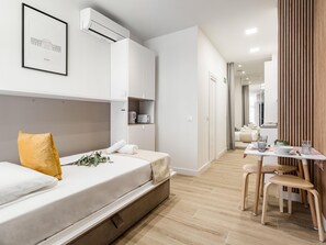 Une chambre d’hôtel moderne, équipée d’un lit, d’un bureau et d’une chaise. Un tableau encadré orne le mur, il y a une fenêtre et une porte qui donne sur une autre pièce.