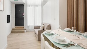 Un espace repas moderne avec une table en verre pour quatre personnes, un canapé avec des coussins et un escalier menant à une porte sombre portant l’inscription « SORTIE ».