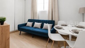 A modern living room with a blue sofa, white chairs, a dining table set for four, and a window with curtains. - Villa de Vallecas I - One-bedroom apartment (Madrid)