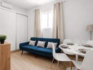 Une pièce moderne avec un canapé bleu, des chaises blanches, une table à manger dressée avec des assiettes et des verres, une plante en pot et une fenêtre avec des rideaux.