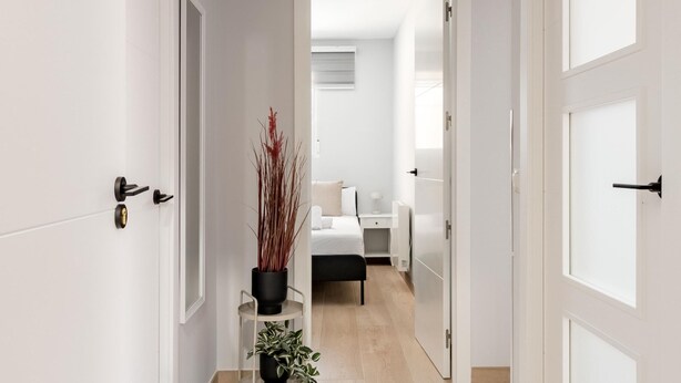 Un pasillo con puertas blancas, un pequeño soporte para planta y una cama al fondo.
