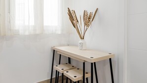 Une pièce au décor minimaliste, avec une table de couleur claire et deux chaises, une fenêtre ornée de rideaux légers et un vase contenant des plantes séchées, posé sur la table.
