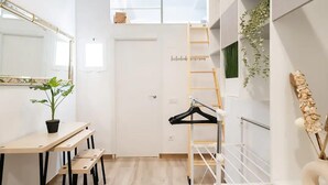 Quarto compacto e moderno com mezanino, uma escrivaninha com cadeiras, um cabideiro e um vaso de plantas.