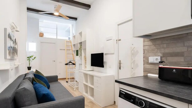 Uma cozinha compacta com máquina de lavar, um sofá cinzento e uma televisão.