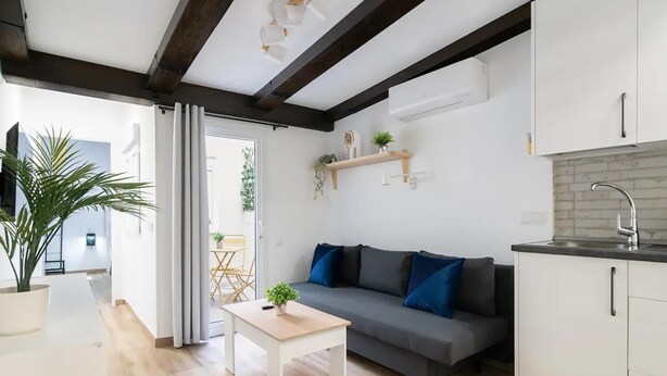 Una sala de estar moderna con un sofá gris, una mesita blanca y una cocina con armarios blancos y una encimera negra.