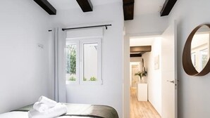 Una habitación de hotel moderna con cama, ventana con cortinas, piso de madera y un pasillo que conduce a otra habitación.