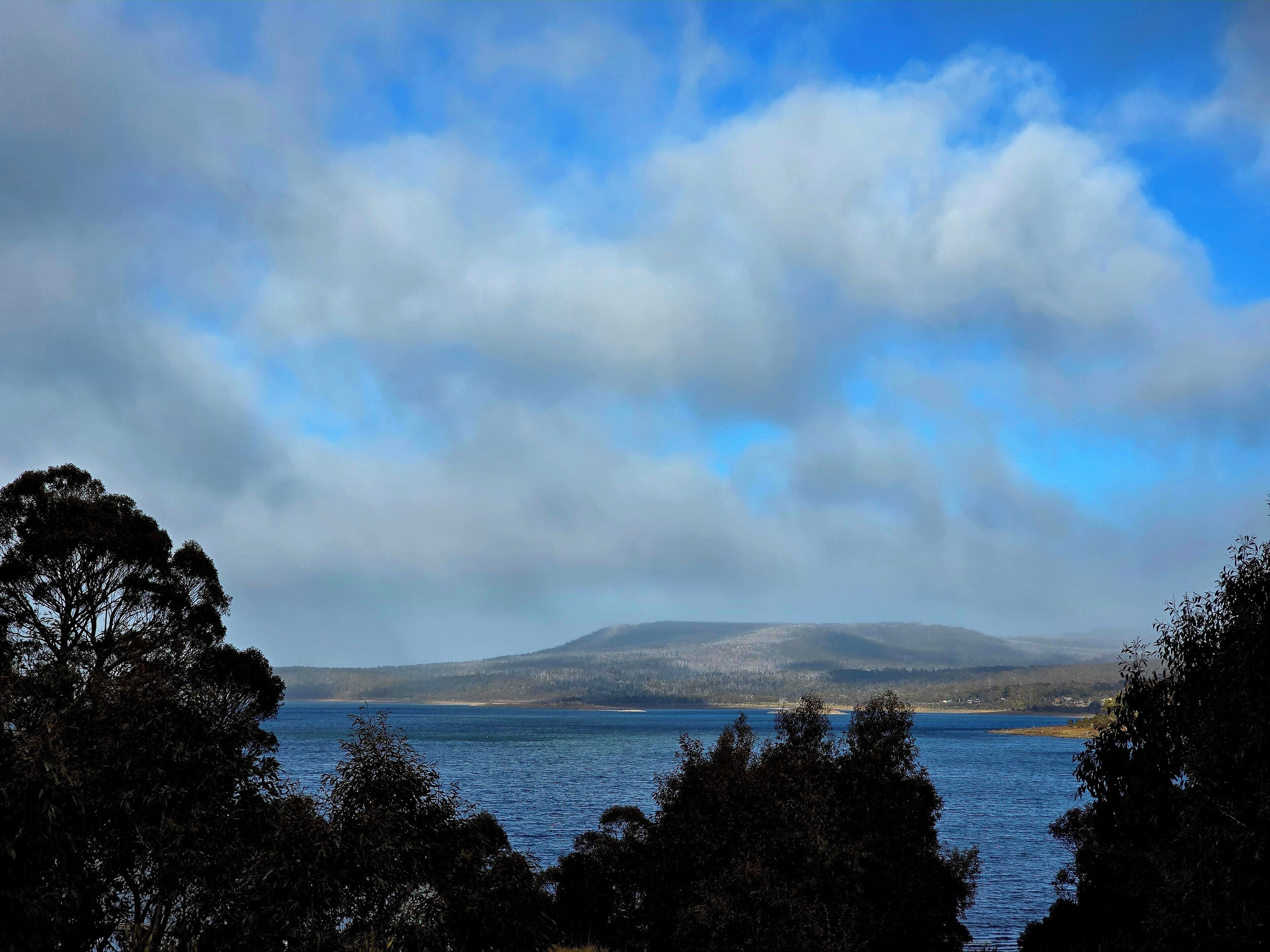 A coastal landscape with a body of water, trees, and a distant mountain range under a partly cloudy sky.