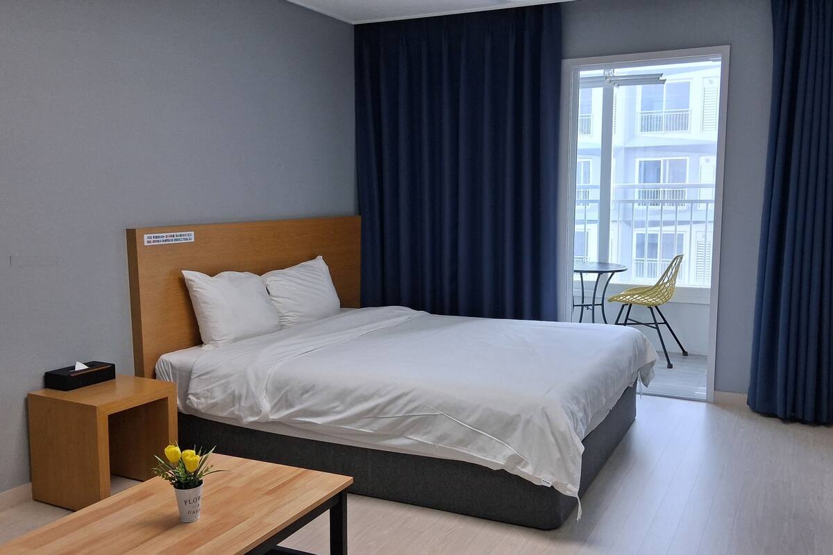 A hotel room with a bed, a wooden nightstand, a small table with a vase of flowers, a chair, and a window with curtains.