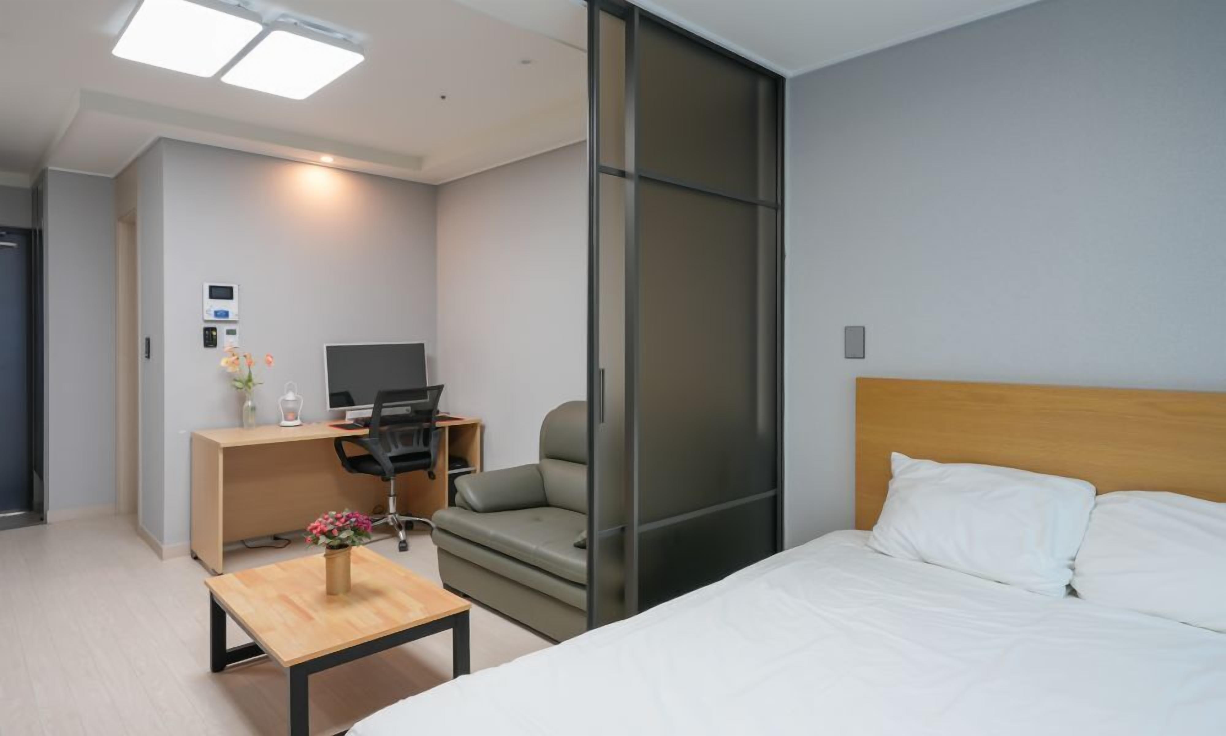 A hotel room with a bed, a desk with a computer, a chair, a small table with flowers, and a sliding door.
