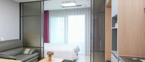 Une chambre d’hôtel moderne avec un canapé, une petite table en bois, un lit avec du linge de maison blanc, une fenêtre avec des rideaux légers et un lave-linge.