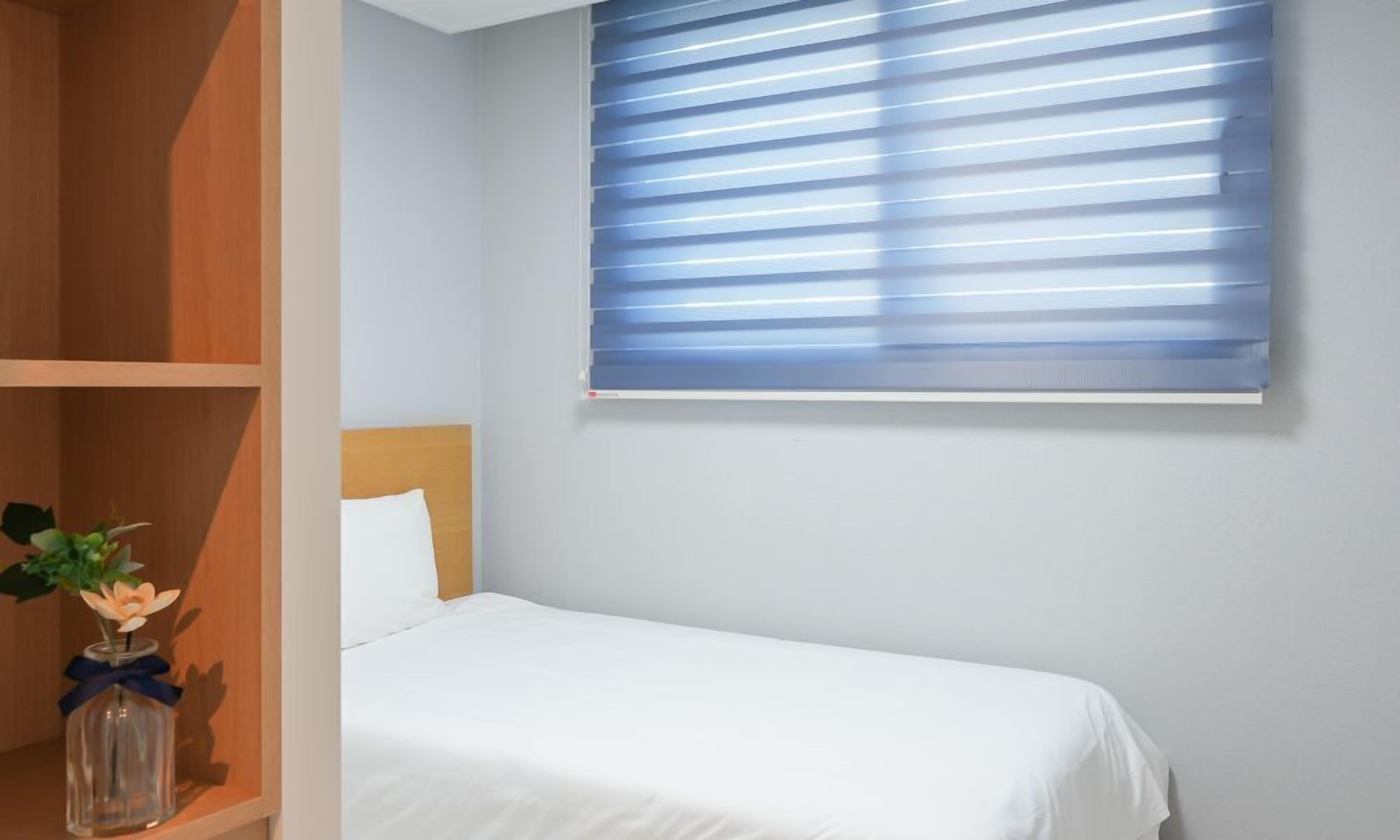 A single bed with white bedding, a wooden headboard, and a window with blue blinds.