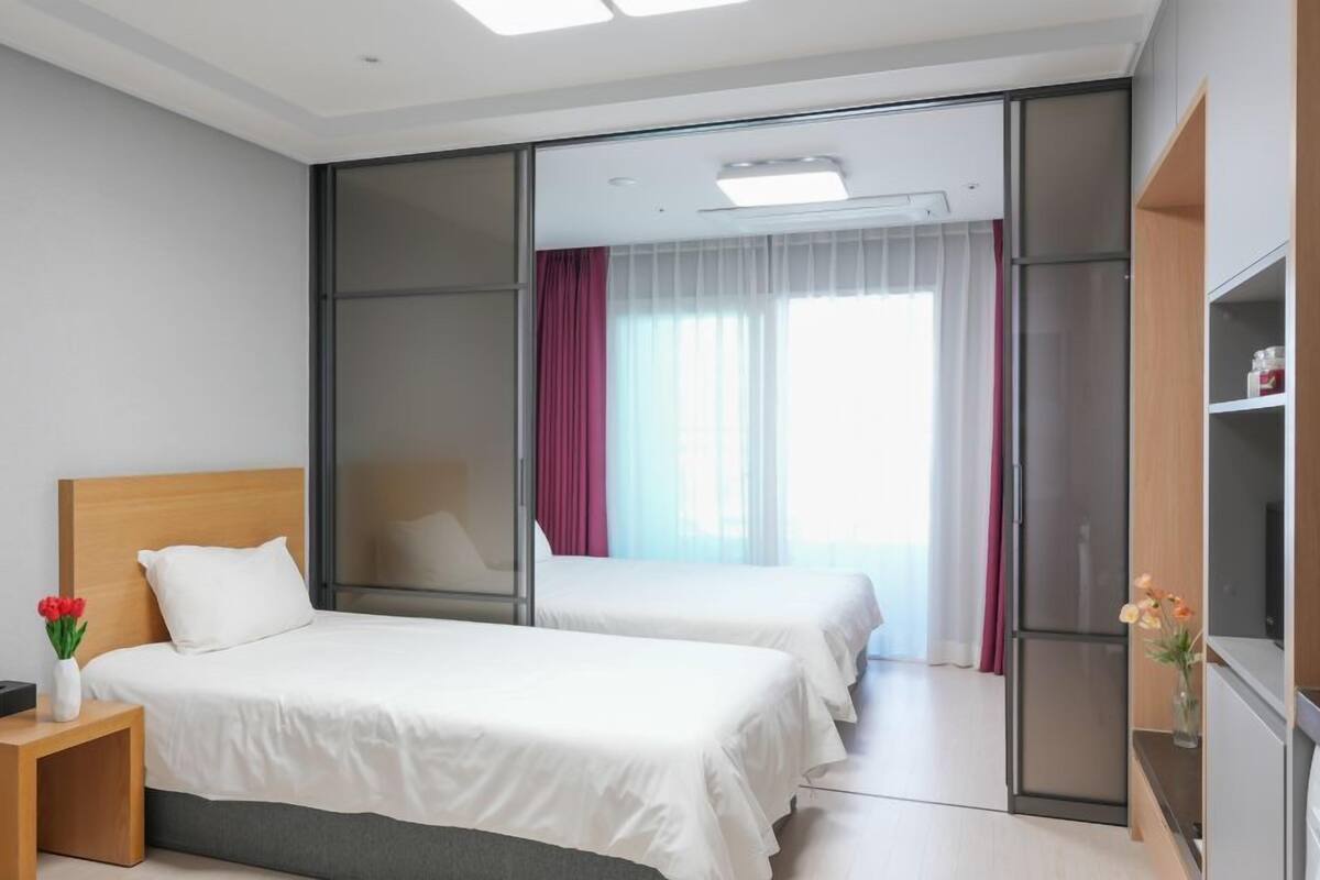 A hotel room with two beds, a desk, a wardrobe, and a large window with curtains.