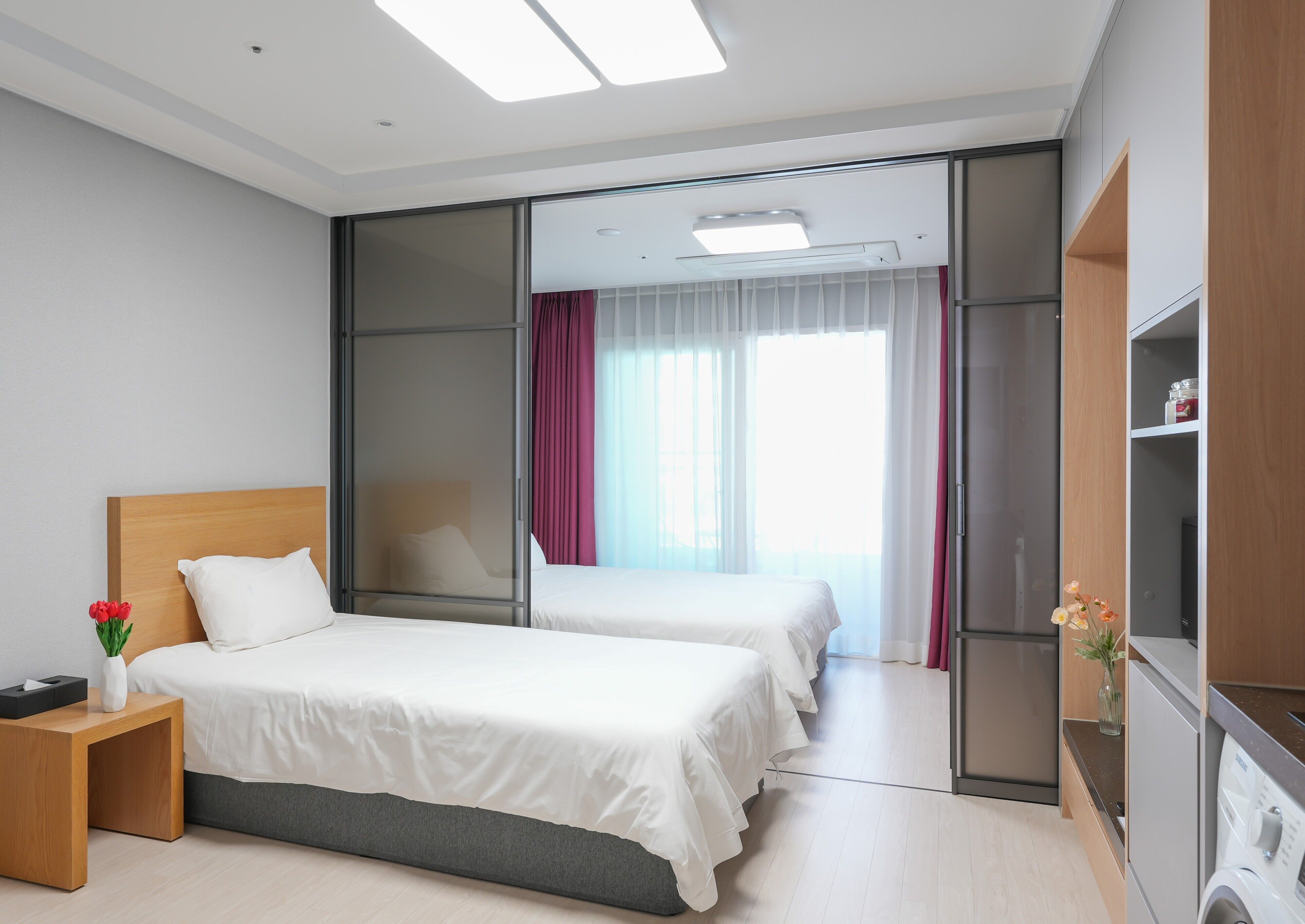 Habitación de hotel con dos camas, una mesita de noche de madera, una pequeña mesa con un florero y una lavadora, además de un ventanal corredero que da a un balcón.