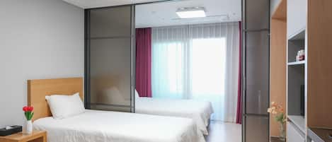 Habitación de hotel con dos camas, una mesita de noche de madera, una pequeña mesa con un florero y una lavadora, además de un ventanal corredero que da a un balcón.