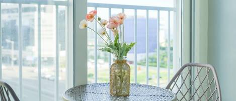 Una mesa redonda con un florero tejido que contiene flores, ubicada cerca de una ventana con vista a una calle y edificios.