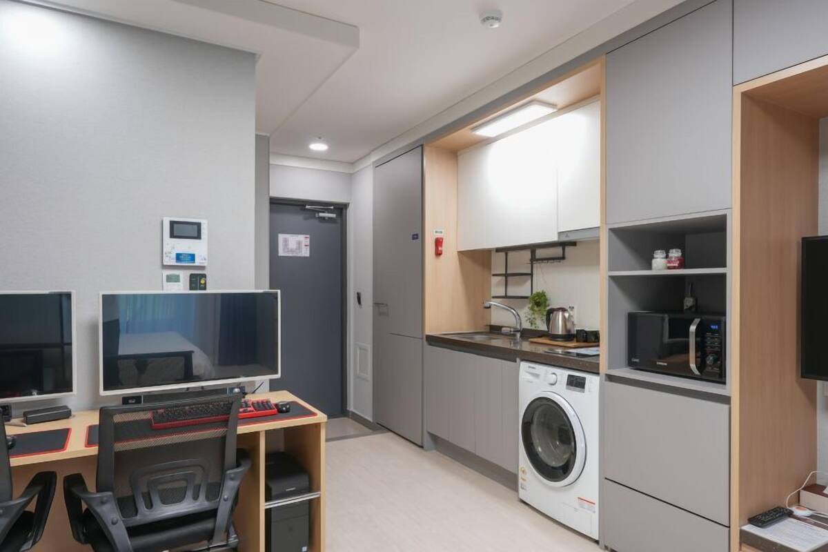 A modern hotel room with a desk, two monitors, a washing machine, and a kitchenette.