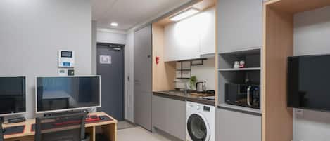 A modern hotel room with a desk, two monitors, a washing machine, and a kitchenette.