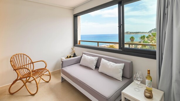 A room with a grey sofa, a wicker chair, a small table with a bottle and glass, and a large window with a view of the sea and hills. - Bonita Stay B Sun: Great views, new, fiber, airconditioning, pool, BBQ & parking (Bahia De Javea (Urbanizacion))