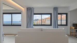 A modern bedroom with a white sofa, two windows with curtains, a desk with a laptop, and a view of buildings and the sea. - Bonita Stay B Sun: Great views, new, fiber, airconditioning, pool, BBQ & parking (Bahia De Javea (Urbanizacion))