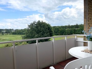 A balcony with a white railing, a table with a vase of flowers, and a view of a green landscape with trees and houses in the distance.