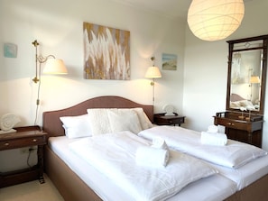 Un lit double avec du linge de lit blanc, deux tables de chevet avec des lampes, un grand miroir et des œuvres d’art accrochées au mur.