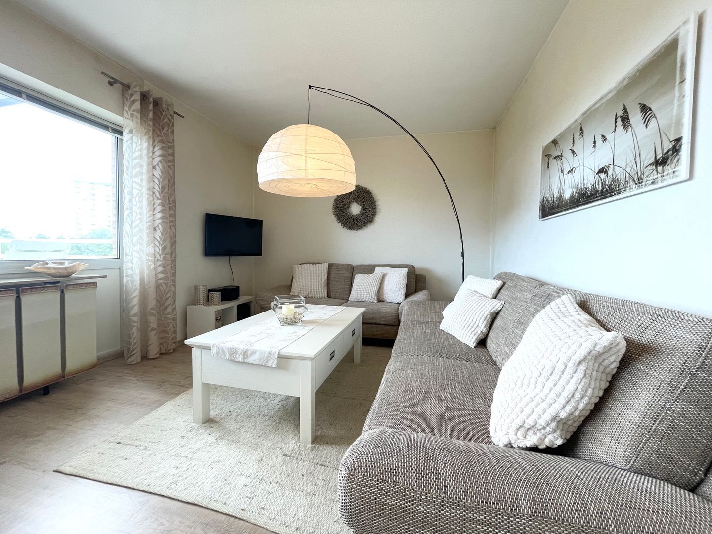 Ein modernes Wohnzimmer mit einer Couch, einem Couchtisch, einem Fernseher und einem großen Fenster mit Vorhängen.