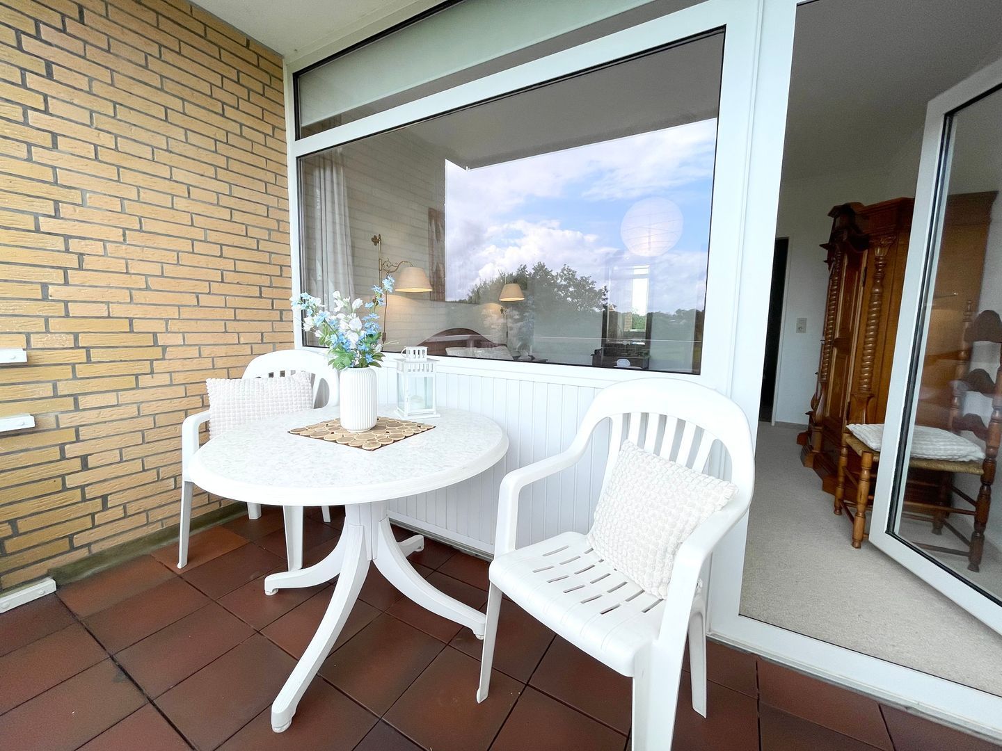 Eine Terrasse mit einem runden Tisch und zwei weißen Stühlen, einer Ziegelwand und einem großen Fenster mit Blick auf Himmel und Bäume.