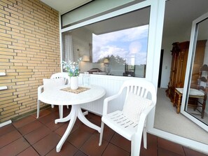Une terrasse avec une table ronde et deux chaises blanches, un mur de briques et une grande fenêtre donnant sur le ciel et les arbres.