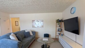 A living room with a sofa, a coffee table, a TV, a clock, and a bookshelf. - In Town 2BR: Dedicated Workspace, Free Wifi, Full Kitchen, and Laundry Access. (Fairbanks)