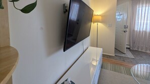 A modern living room with a flat-screen TV mounted on a white wall, a standing lamp, a door with a number, and a plant. - In Town 2BR: Dedicated Workspace, Free Wifi, Full Kitchen, and Laundry Access. (Fairbanks)
