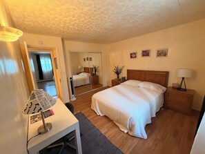 A bedroom with a bed, bedside tables, a desk with a lamp, a mirror, and a door leading to another room.