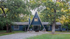 A modern house with a triangular roof, a patio with outdoor seating, and a large tree in the foreground.