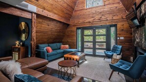 A cozy loft-style room with a wooden ceiling, a large window, a sofa, a coffee table, and a fireplace.