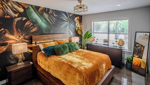 A bedroom with a large bed, a nightstand, a lamp, a plant, and a window with blinds.