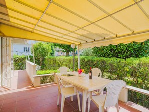 Eine überdachte Terrasse mit Tisch und Stühlen, einer Weinflasche und Blick auf Grünflächen und Gebäude.