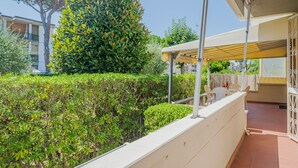 Ein Balkon mit weißer Brüstung, einer überdachten Sitzgelegenheit und Blick auf einen ordentlich geschnittenen Heckenstreifen und einen hohen Baum.