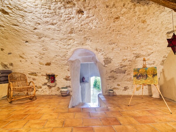 An interior with a rustic stone ceiling, a doorway leading outside, a wicker chair, and an easel with a painting.