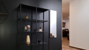 Una sala de estar moderna con una estantería negra sobre una pared oscura, suelo de madera y una cocina visible al fondo.