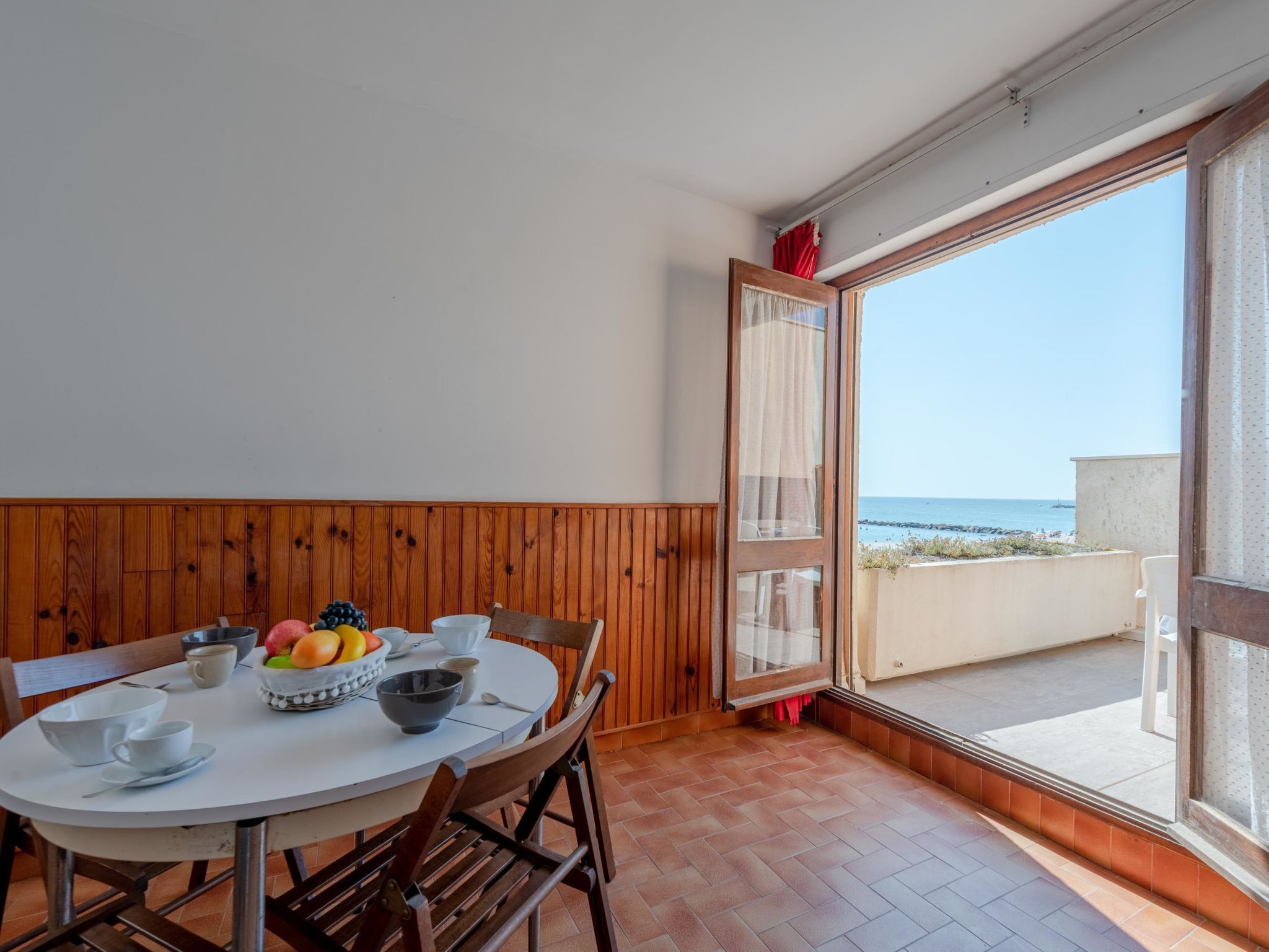 Een eethoek met een ronde tafel, stoelen en een schaal met fruit. Een groot raam biedt uitzicht op de zee.
