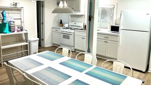 A modern kitchen with white cabinets, a stove, and a refrigerator. There is a dining table with chairs and a hanging light fixture. - HOT TUB, beach access, 2-bdrm cottage just outside Port Austin with AC & WiFi #1 (Port Austin)