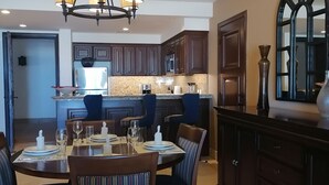 Uma área de refeições com uma mesa redonda posta para quatro pessoas, rodeada por cadeiras estofadas. Uma cozinha com armários de madeira escura e bancada de granito é visível ao fundo.