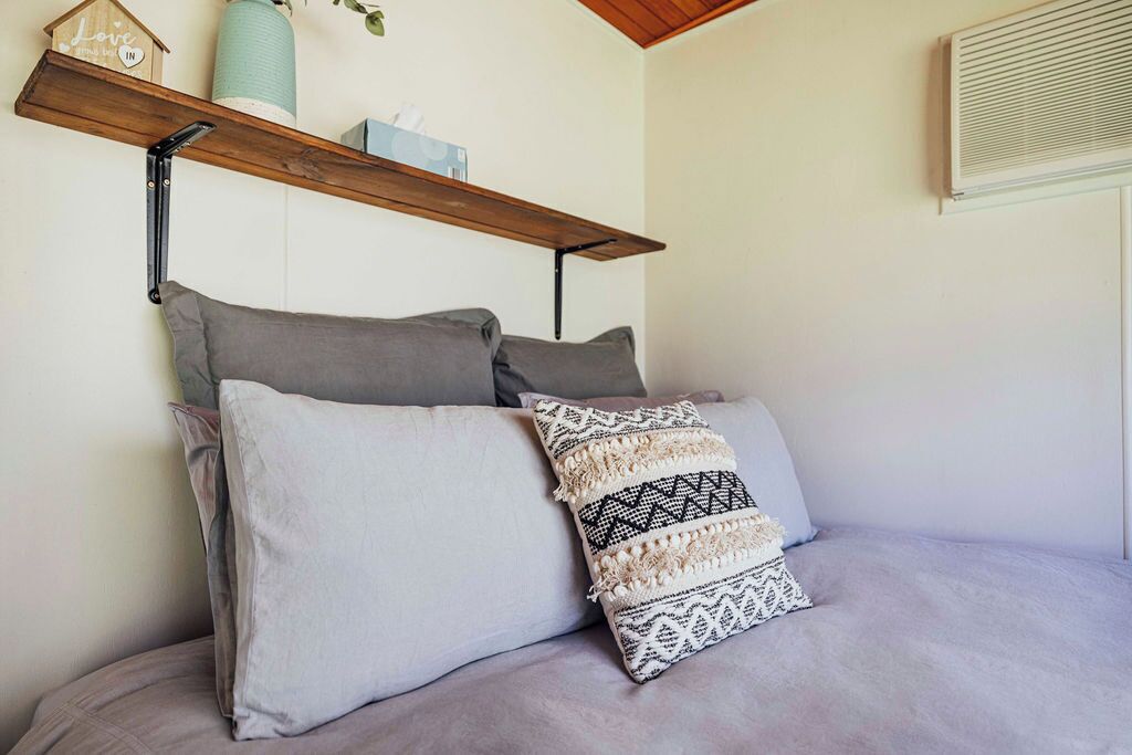 A bed with grey bedding and decorative pillows, a wooden shelf with a small plant, and a wall-mounted air vent.