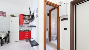 Une cuisine de taille réduite, équipée d’un réfrigérateur, de placards rouges, d’un plan de travail noir et d’une porte donnant sur une salle de bain.