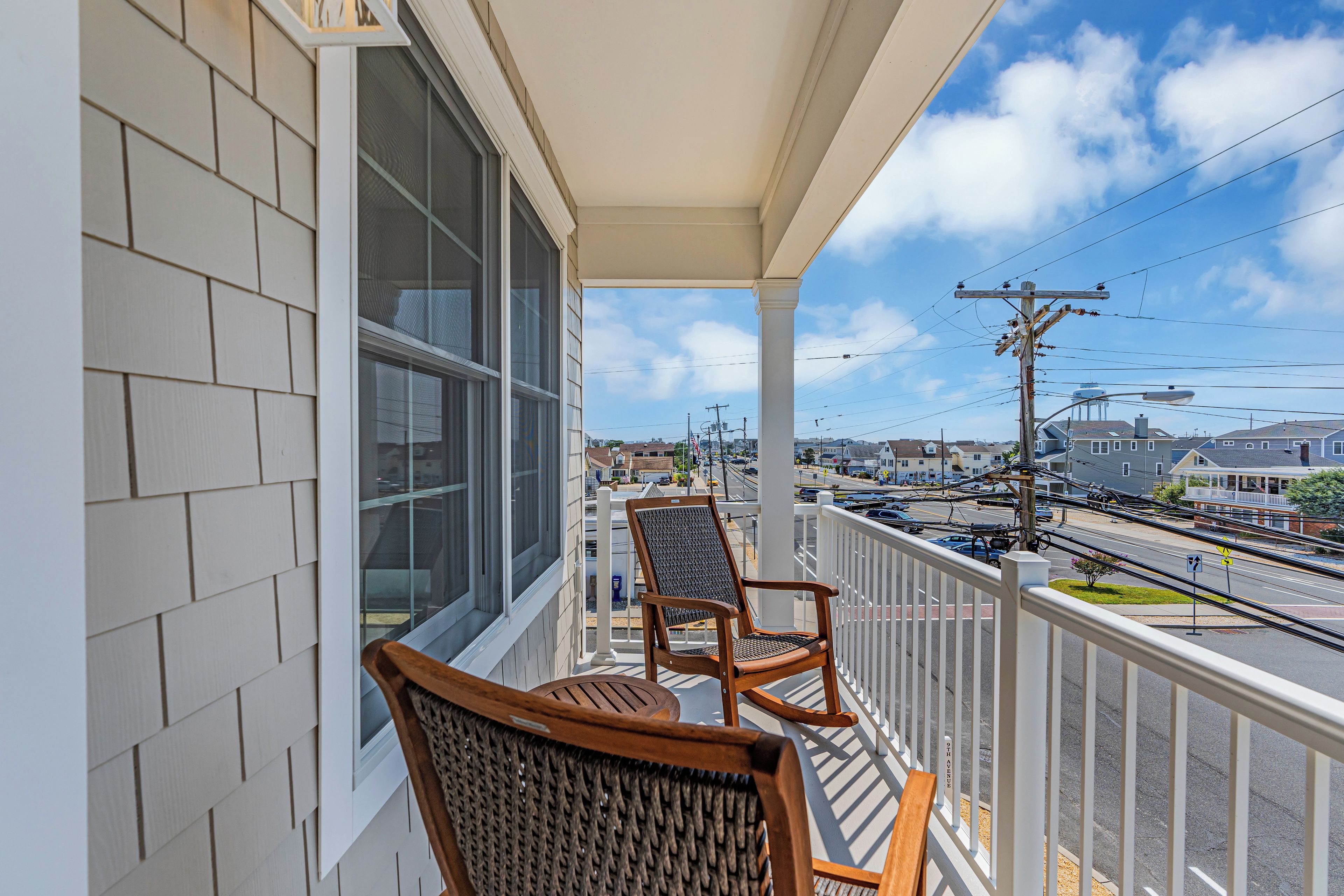 A balcony with two wicker chairs and a wooden table, overlooking a street with buildings and power lines.