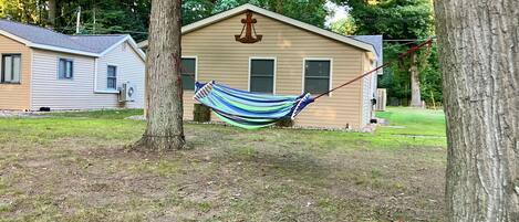 A hammock tied between two trees in a grassy area with a house in the background.