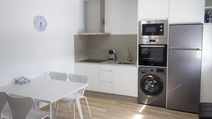 A compact kitchen with a microwave, oven, and washing machine, a dining table with chairs, and a clock on the wall.
